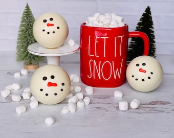 Puede incluir: Tres bombas de chocolate caliente de chocolate blanco con forma de muñeco de nieve con ojos negros y nariz roja, junto a una taza roja con malvaviscos blancos y el texto "Let it Snow".