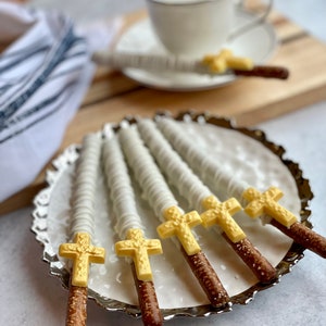 May include: A plate of six pretzel rods dipped in white chocolate and topped with gold cross designs. The pretzels are arranged in a fan shape on a silver plate.