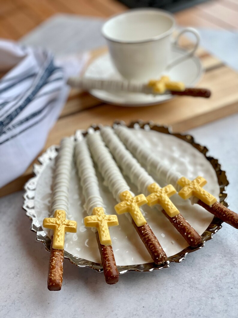 May include: A plate of six pretzel rods dipped in white chocolate and topped with gold cross designs. The pretzels are arranged in a fan shape on a silver plate.