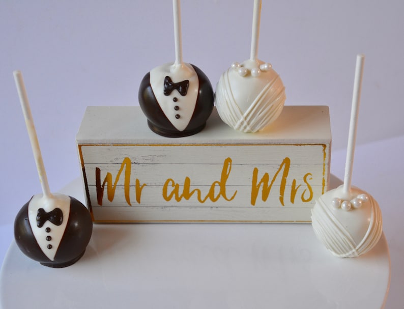 May include: Four cake pops decorated to look like a bride and groom. The cake pops are on a white cake stand with a wooden sign that says "Mr. and Mrs."