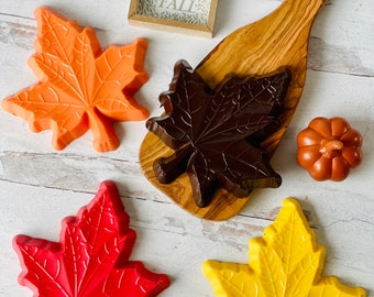 May include: Four fall-themed maple leaf-shaped chocolates in orange, red, yellow, and brown. The chocolates are arranged on a white wooden surface with a small wooden sign that says "Hello Fall".