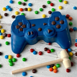 May include: A blue chocolate video game controller with brown buttons and a wooden mallet. The controller is surrounded by colorful candy.