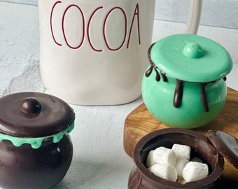 May include: Three chocolate pots with lids. One pot is green and the other two are brown. One of the brown pots is filled with white marshmallows. A white mug with the word "COCOA" in red lettering is in the background.