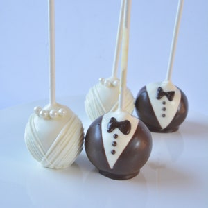 May include: Four cake pops decorated to look like a bride and groom. Two cake pops are white with pearl accents and two cake pops are brown with white accents.