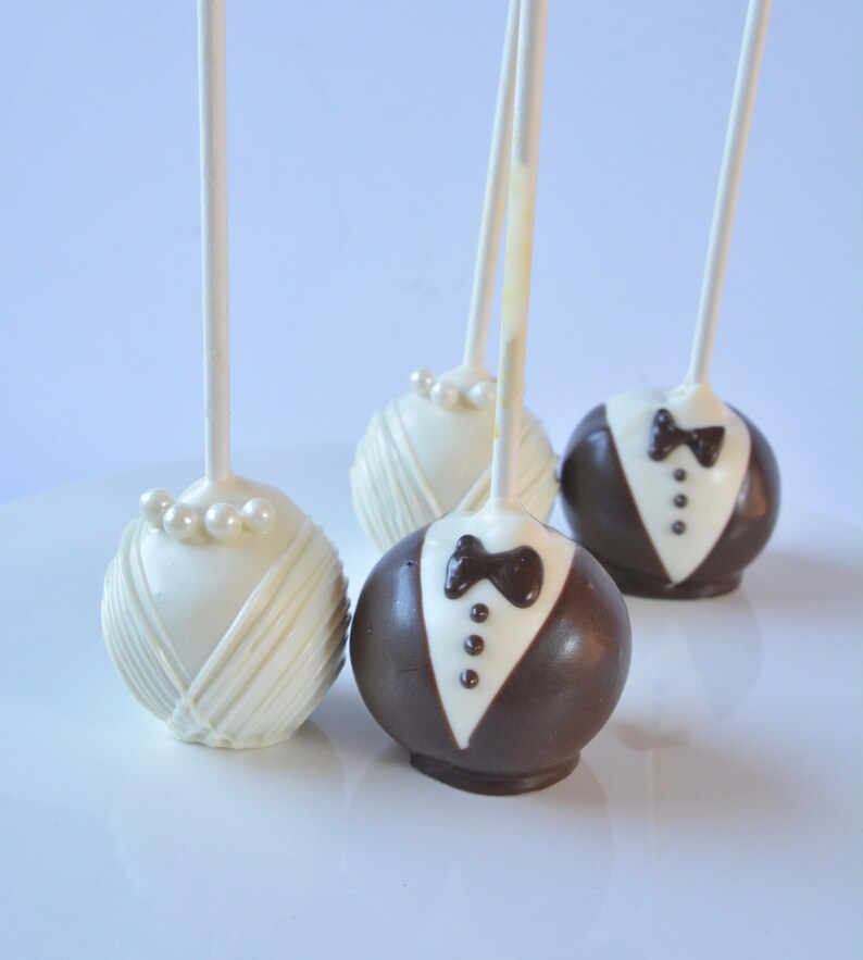 May include: Four cake pops decorated to look like a bride and groom. Two cake pops are white with pearl accents and two cake pops are brown with white accents.