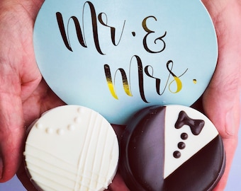 May include: Two decorated cookies, one white with a striped design and the other black and white with a tuxedo design, are held in a hand with a blue and gold 'Mr. & Mrs.' sign behind them.