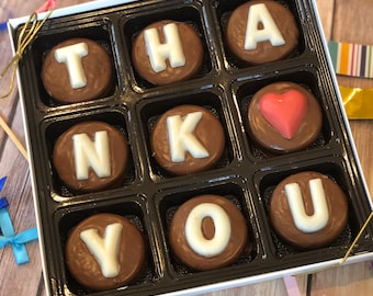 May include: A white box filled with chocolate covered cookies that spell out "THANK YOU". The cookies are arranged in a 3x4 grid. The "A" and "U" are in the top right and bottom right corners, respectively. The "K" is in the middle row, second from the right. The "H" is in the top row, second from the right. The "N" is in the middle row, second from the left. The "T" is in the top row, second from the left. The "Y" is in the bottom row, second from the left. The "O" is in the bottom row, second from the right. There is a heart-shaped cookie in the middle row, third from the left.
