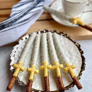 May include: Six white chocolate covered pretzel rods with gold cross toppers on a silver platter. The pretzels are arranged in a fan shape.