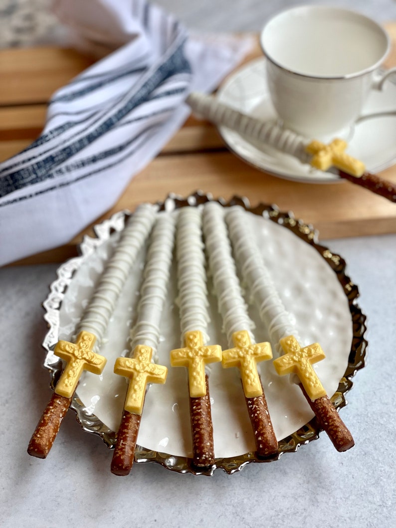 May include: Six white chocolate covered pretzel rods with gold cross toppers on a silver platter. The pretzels are arranged in a fan shape.