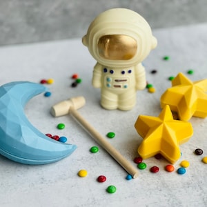 May include: An astronaut figurine, a blue crescent moon, and two yellow star-shaped objects are arranged on a light gray surface. A small wooden mallet and colorful candies are also present. The astronaut has a gold-colored visor.