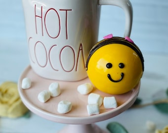 Puede incluir: Una taza de cerámica blanca con las palabras "HOT COCOA" impresas en rojo, llena de malvaviscos, junto a un cake pop amarillo con cara sonriente y lazos rosas en un soporte para pasteles rosa.