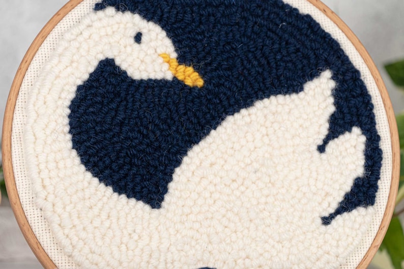 K&ouml;nnte beinhalten: Eine wei&szlig;e und marineblaue Nadelfilz-Stickerei eines Schwans mit gelbem Schnabel. Der Schwan ist zu einem Kreis zusammengerollt, mit dem Kopf unter dem Fl&uuml;gel. Die Stickerei ist in einem Holzrahmen eingefasst.