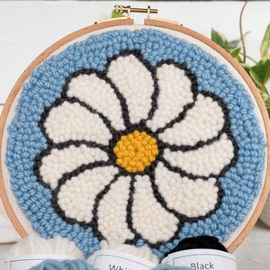 Puede incluir: Un aro de bordado de punto de aguja azul y blanco con un dise&ntilde;o de margarita. La margarita es blanca con un centro amarillo y contornos negros. El aro es de madera y tiene un anillo de metal dorado.