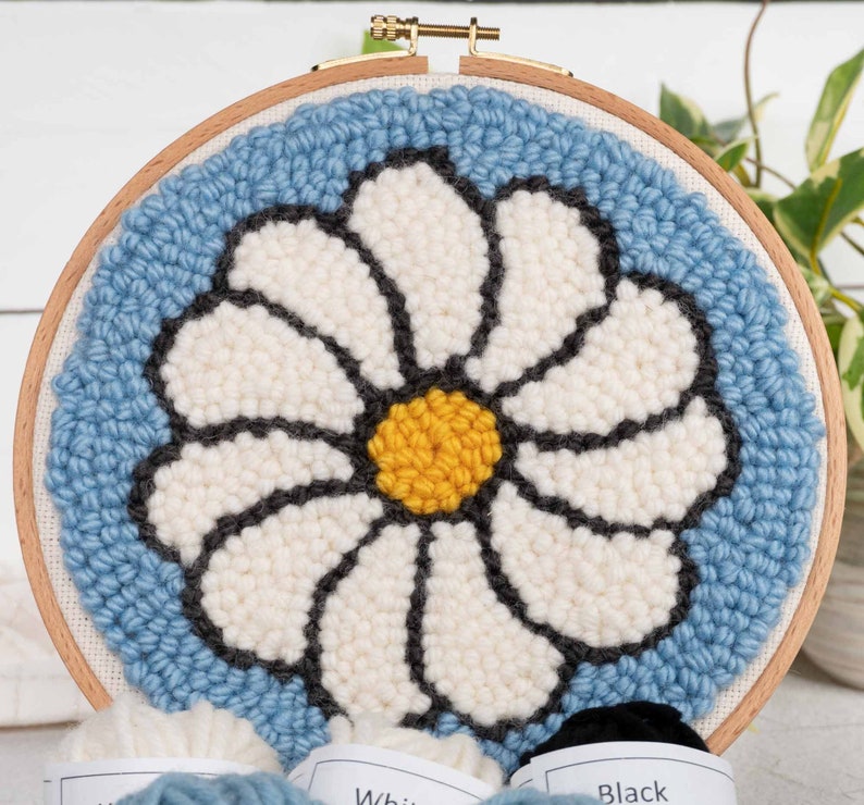 Puede incluir: Un aro de bordado de punto de aguja azul y blanco con un dise&ntilde;o de margarita. La margarita es blanca con un centro amarillo y contornos negros. El aro es de madera y tiene un anillo de metal dorado.