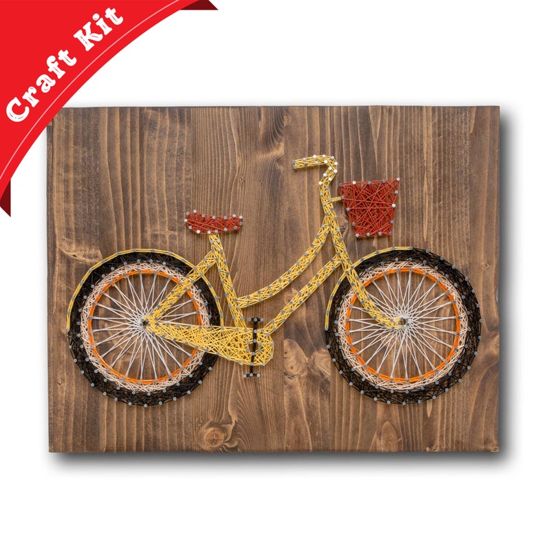 Puede incluir: Una bicicleta amarilla con una cesta en el manillar est&aacute; hecha con cuerda envuelta alrededor de clavos en una tabla de madera. La bicicleta es un kit de arte de cuerda.