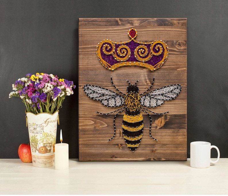 May include: A wooden wall art piece featuring a bee with a crown above it. The bee is made of yellow, black, and white string art. The crown is made of purple and gold string art. The background is a dark brown wood.