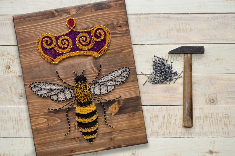 May include: A wooden board with a bee and a crown made of string art. The bee is yellow and black, and the crown is purple and gold. There are nails and a hammer on a wooden surface next to the board.