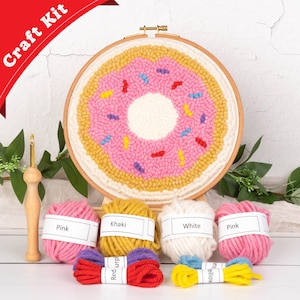 May include: A craft kit featuring a donut design. The kit includes a wooden embroidery hoop with a donut design in pink, yellow, and white yarn, along with several skeins of yarn in pink, khaki, and white. The text "Craft Kit" is visible.