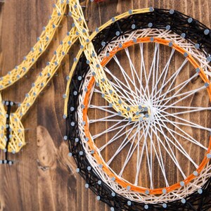 Puede incluir: Arte de hilo de una bicicleta amarilla con ruedas negras y naranjas sobre un fondo de madera.