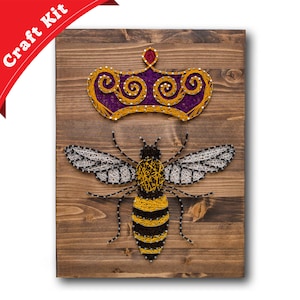 May include: A string art craft kit featuring a bee with a crown on a wooden background. The bee is made of yellow, black, and white string, and the crown is made of purple and yellow string.