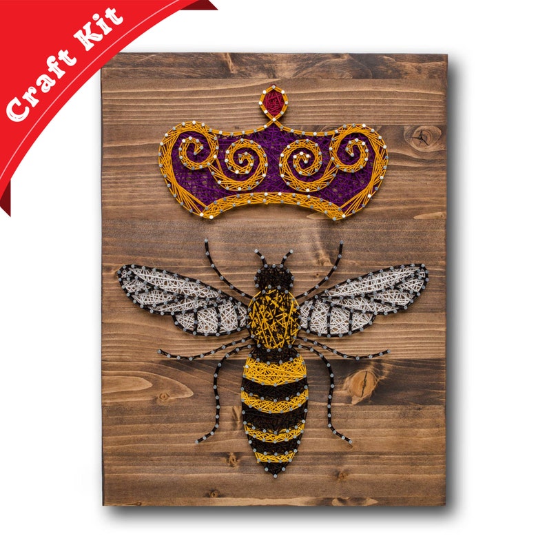 May include: A string art craft kit featuring a bee with a crown on a wooden background. The bee is made of yellow, black, and white string, and the crown is made of purple and yellow string.