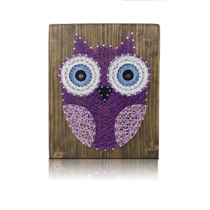 May include: A handcrafted string art owl on a wooden board. The owl features purple, white, and light blue string, with a dark brown wooden background. The owl's eyes are detailed with concentric circles of white and blue string.