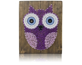 May include: A handcrafted string art owl on a wooden board. The owl features purple, white, and light blue string, with a dark brown wooden background. The owl's eyes are detailed with concentric circles of white and blue string.