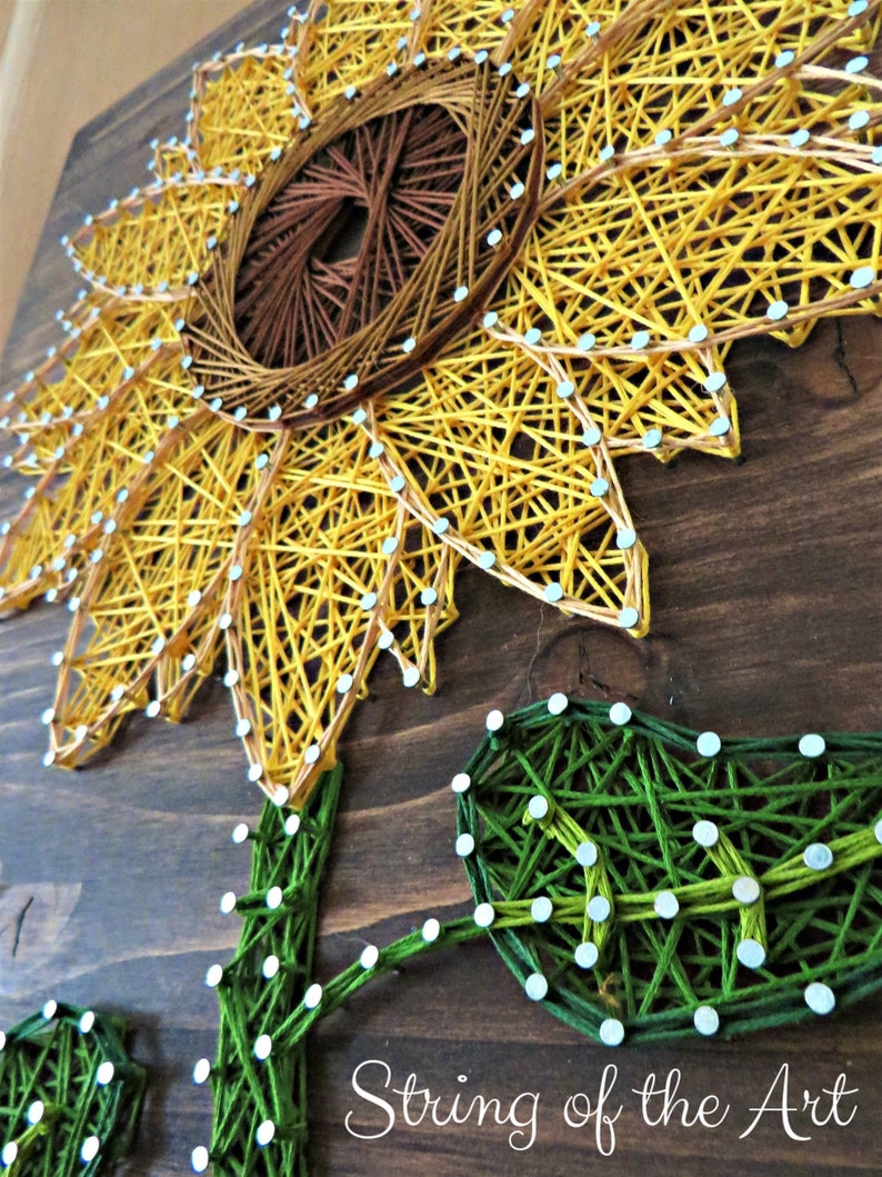 Pu&ograve; includere: Arte di filo di un girasole con un centro marrone, petali gialli e foglie verdi. L'arte &egrave; realizzata con lo spago avvolto attorno a chiodi martellati su una tavola di legno. Il testo "String of the Art" &egrave; visibile nella parte inferiore dell'immagine.