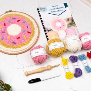 May include: A donut punch needle kit with a pink frosted donut design. The kit includes yarn in various colors, a punch needle tool, a marker, and instructions. The finished piece is in a wooden hoop.