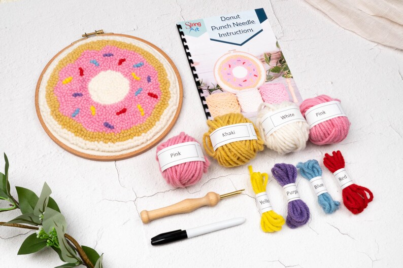 May include: A donut punch needle kit with a pink frosted donut design. The kit includes yarn in various colors, a punch needle tool, a marker, and instructions. The finished piece is in a wooden hoop.