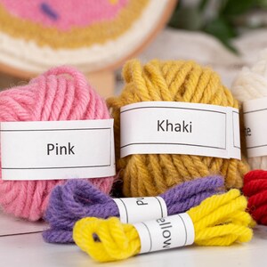 May include: A collection of colorful yarn balls and embroidery floss in pink, khaki, white, purple, yellow, red, and blue. Each yarn ball has a label with its color name. A wooden punch needle tool and a partially completed embroidery hoop are also visible.