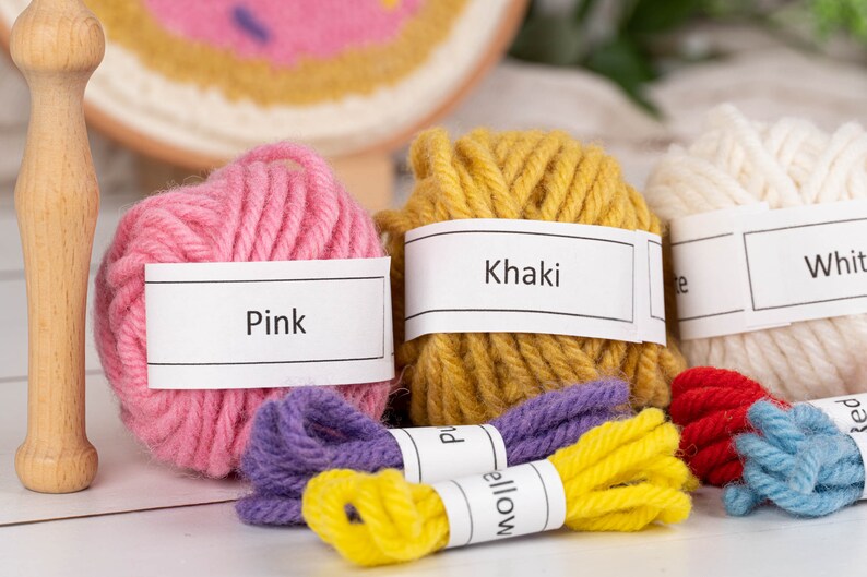 May include: A collection of colorful yarn balls and embroidery floss in pink, khaki, white, purple, yellow, red, and blue. Each yarn ball has a label with its color name. A wooden punch needle tool and a partially completed embroidery hoop are also visible.
