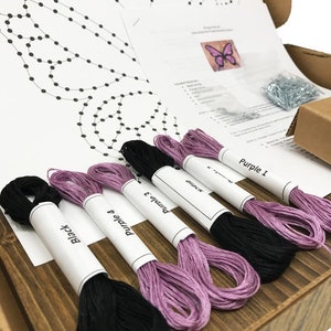 May include: A craft kit for string art featuring a butterfly design. The kit includes a printed pattern, embroidery floss in black and purple, and tacks. The floss is labeled "Black", "Purple 4", "Purple 3", "Purple 2", and "Purple 1".