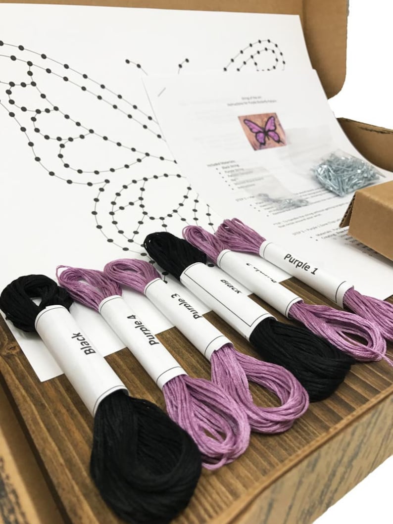 May include: A craft kit for string art featuring a butterfly design. The kit includes a printed pattern, embroidery floss in black and purple, and tacks. The floss is labeled "Black", "Purple 4", "Purple 3", "Purple 2", and "Purple 1".