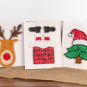 May include: Three string art Christmas decorations on wooden boards. The decorations are a reindeer, Santa Claus stuck in a chimney, and a Christmas tree.