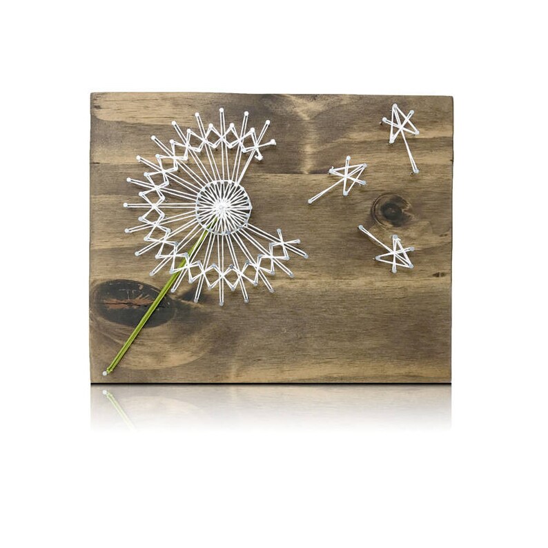 May include: White string art of a dandelion with a green stem on a brown wooden background. Four white stars are scattered around the dandelion.