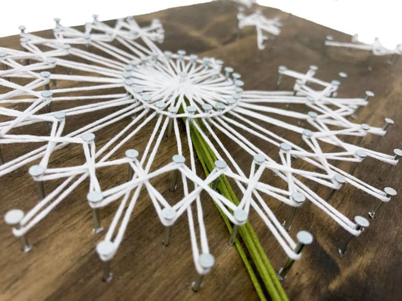 May include: White string art on a wooden background, forming a dandelion shape with a green stem.