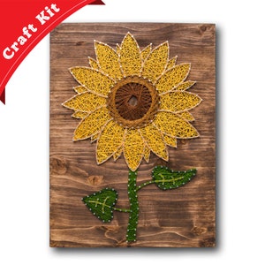 May include: A string art craft kit featuring a sunflower with yellow petals and a brown center. The sunflower is on a wooden background.