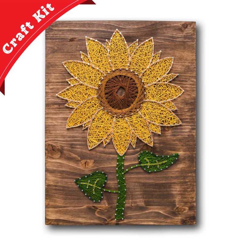 Pu&ograve; includere: Un kit di arte con filo che presenta un girasole con petali gialli e un centro marrone. Il girasole &egrave; su uno sfondo di legno.