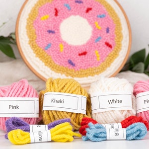 May include: A punch needle kit featuring a donut design. The kit includes a wooden punch needle tool, pink, khaki, white, and pink yarn, and a variety of colorful yarn skeins. The donut design is pink with yellow and colorful sprinkles.