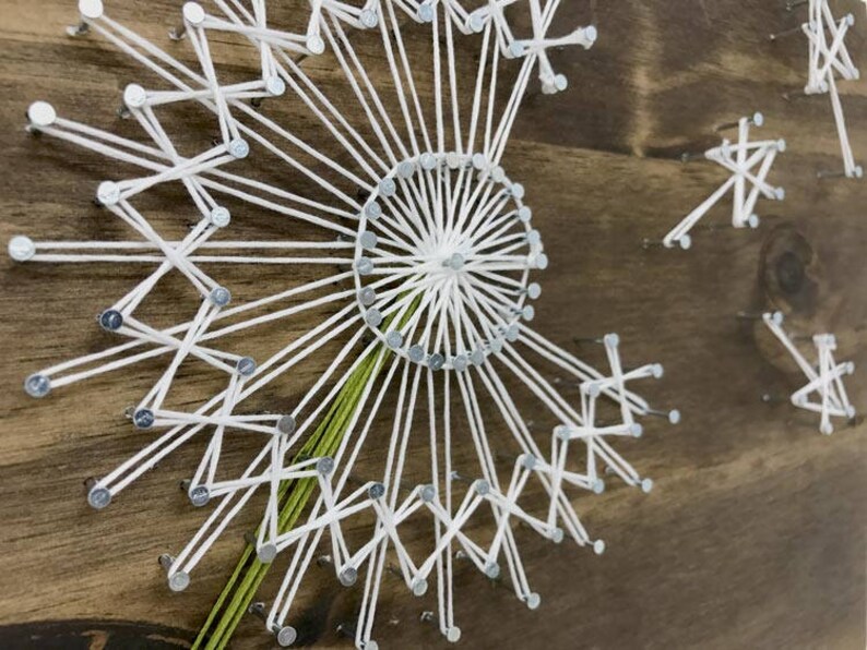 May include: White string art on a wooden background, forming a dandelion shape with green string for the stem.