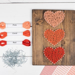 May include: A wooden string art piece featuring three hearts in shades of pink, coral, and red. The hearts are made with string and nails on a dark brown wooden board. Spools of pink, coral, and red thread are also visible.
