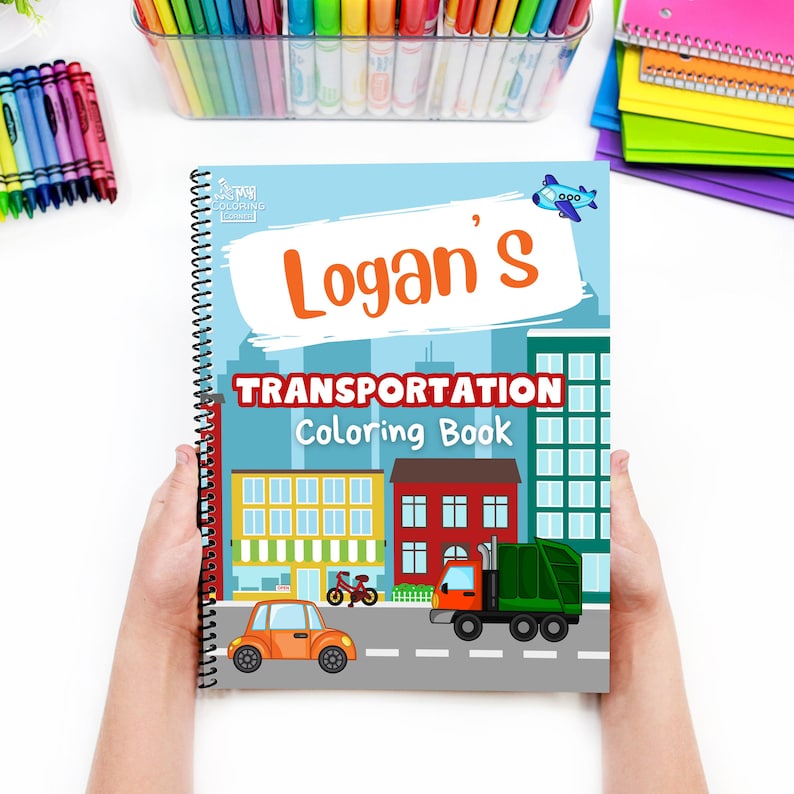 May include: A spiral-bound "Logan's Transportation Coloring Book" with a blue cover featuring illustrations of vehicles and buildings. The title is in red and white, with a cartoon airplane. Crayons and coloured pencils are visible in the background.
