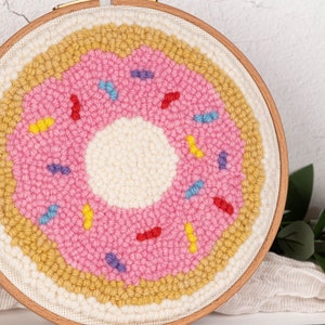 May include: A handmade, circular punch needle art piece depicting a pink donut with colorful sprinkles. The donut is set against a cream-colored background, framed by a wooden embroidery hoop. The artwork is a fun, whimsical home decor item.
