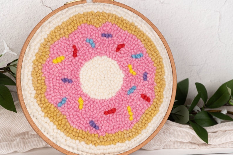 May include: A handmade, circular punch needle art piece depicting a pink donut with colorful sprinkles. The donut is set against a cream-colored background, framed by a wooden embroidery hoop. The artwork is a fun, whimsical home decor item.