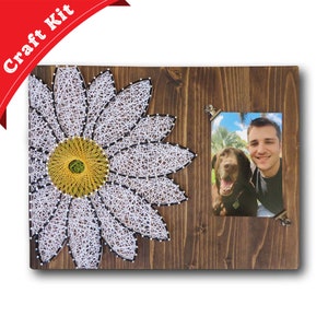 May include: A string art craft kit featuring a white daisy with a yellow center. The kit includes a wooden frame with a photo clip to hold a picture. The picture shows a man and his dog.