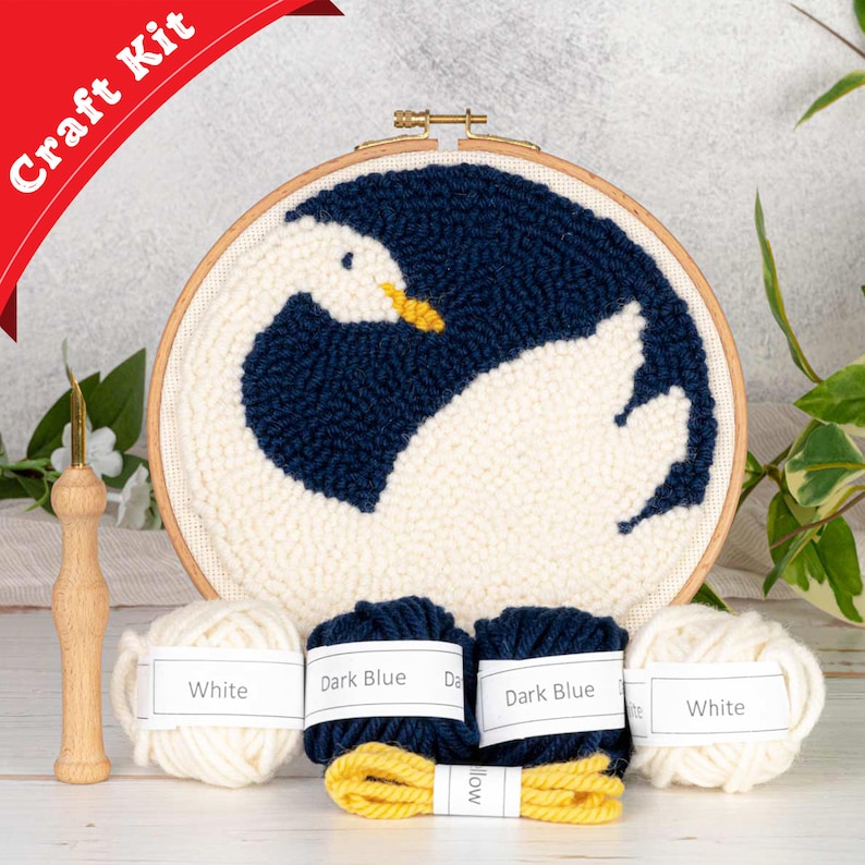 K&ouml;nnte beinhalten: Ein Bastelset zum Herstellen einer Punch-Needle-Stickerei eines wei&szlig;en Schwans mit einem dunkelblauen Fl&uuml;gel. Das Set enth&auml;lt wei&szlig;es, dunkelblaues und gelbes Garn, eine Holz-Nadel und einen Holz-Stickrahmen.