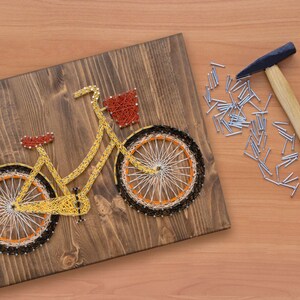 Puede incluir: Una bicicleta amarilla con una cesta est&aacute; hecha con cuerda envuelta alrededor de clavos martillados en una tabla de madera. Un martillo y un mont&oacute;n de clavos est&aacute;n sobre la mesa junto a la tabla.