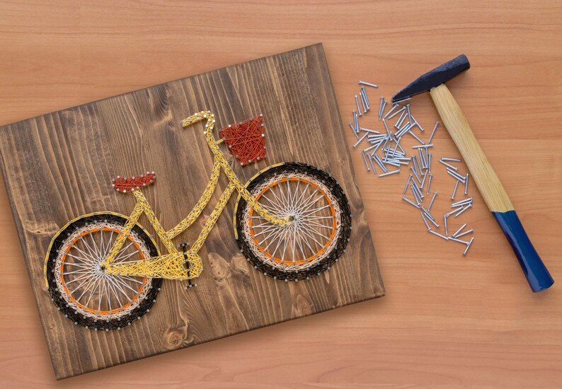 Puede incluir: Una bicicleta amarilla con una cesta est&aacute; hecha con cuerda envuelta alrededor de clavos martillados en una tabla de madera. Un martillo y un mont&oacute;n de clavos est&aacute;n sobre la mesa junto a la tabla.