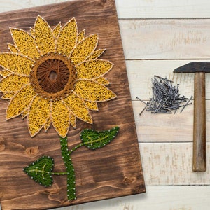 Pu&ograve; includere: Un pannello di legno con un design di girasole in arte con la corda. Il girasole &egrave; realizzato con corda gialla e marrone e ha foglie verdi. Sono mostrati anche un martello e chiodi.
